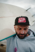 Bearded man in the black Vintage Tiger 5 panel cap and heather grey Neon Sun Wheel hoodie, carrying a surfboard