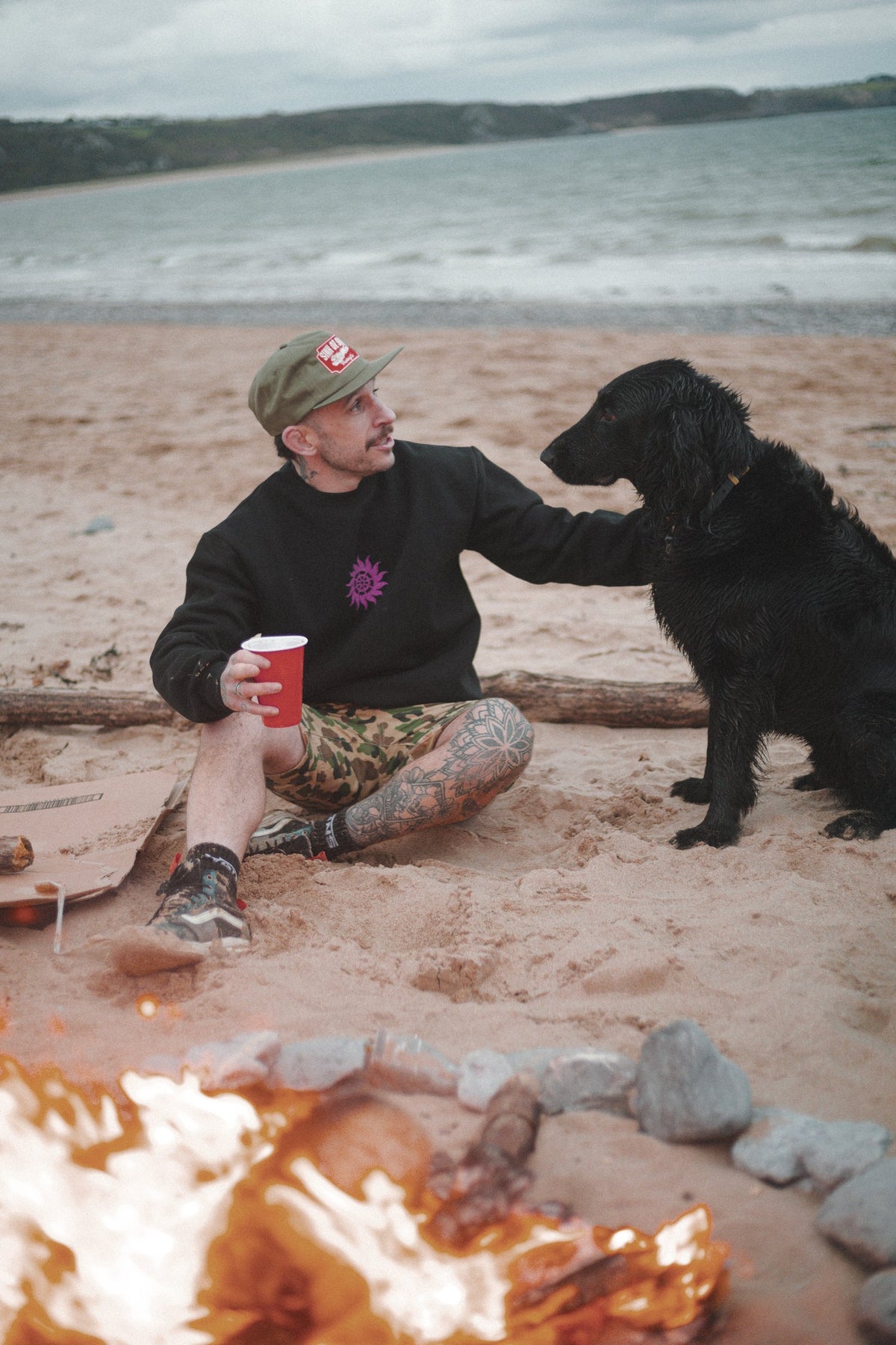 Man in the army green Vintage Tiger cap and black Neon Sun Wheel sweatshirt, stroking a black dog by a beach campfire