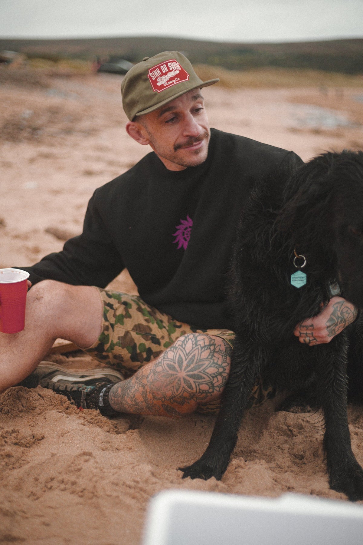 Man in the army green Vintage Tiger 5 panel cap and black Neon Sun Wheel sweatshirt, sitting on the sand with a black dog