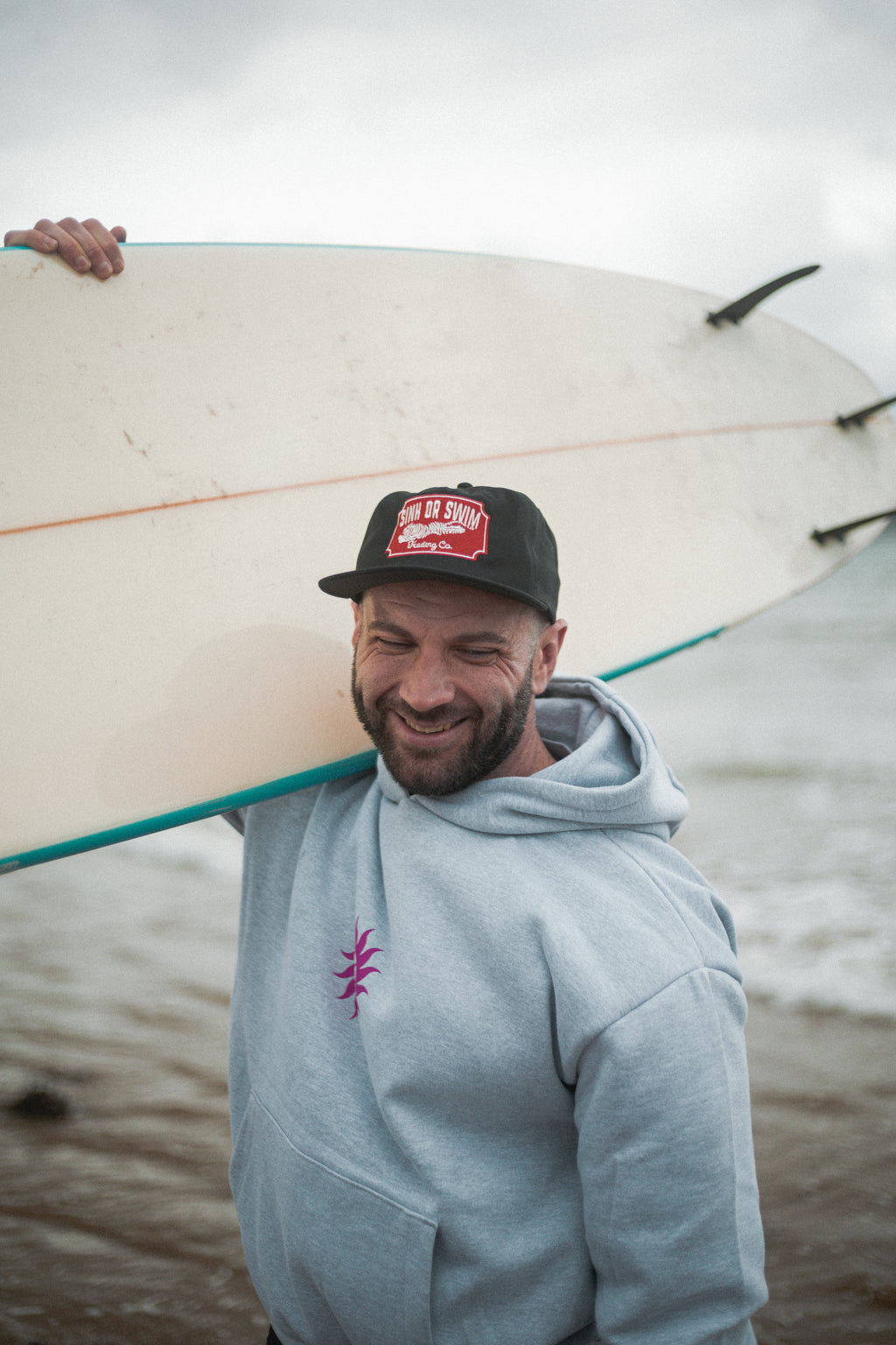 Smiling man carrying a surfboard in the heather grey Neon Sun Wheel heavyweight hoodie and black Vintage Tiger cap