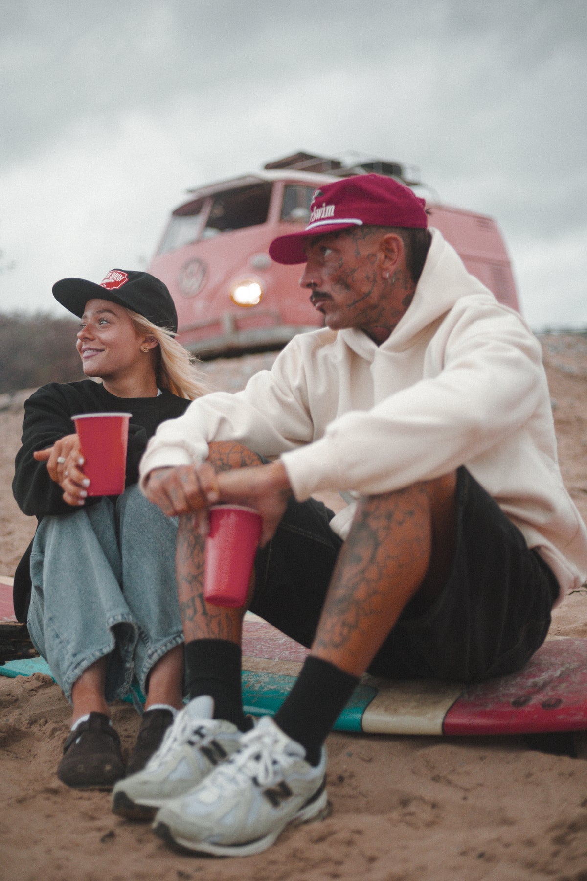 Couple by a pink camper van in a black Neon Sun Wheel sweatshirt and vanilla hoodie, with black Tiger and red Eagle caps