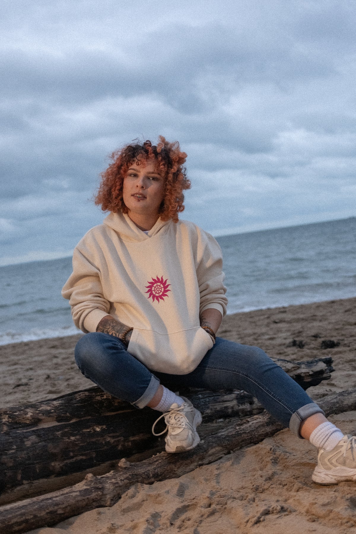 Woman sitting on beach driftwood in the oversized vanilla Neon Sun Wheel heavyweight hoodie with neon pink chest print