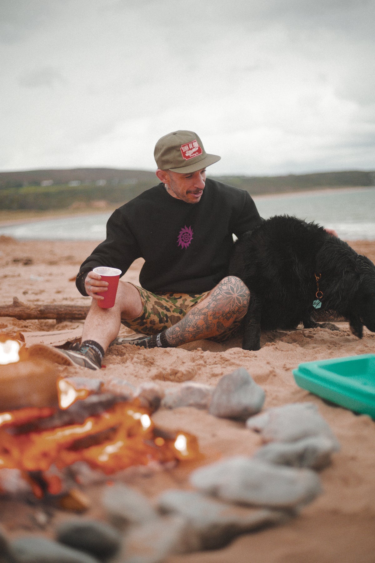 Man in the black Neon Sun Wheel sweatshirt and army green Vintage Tiger cap, by a beach campfire with a black dog