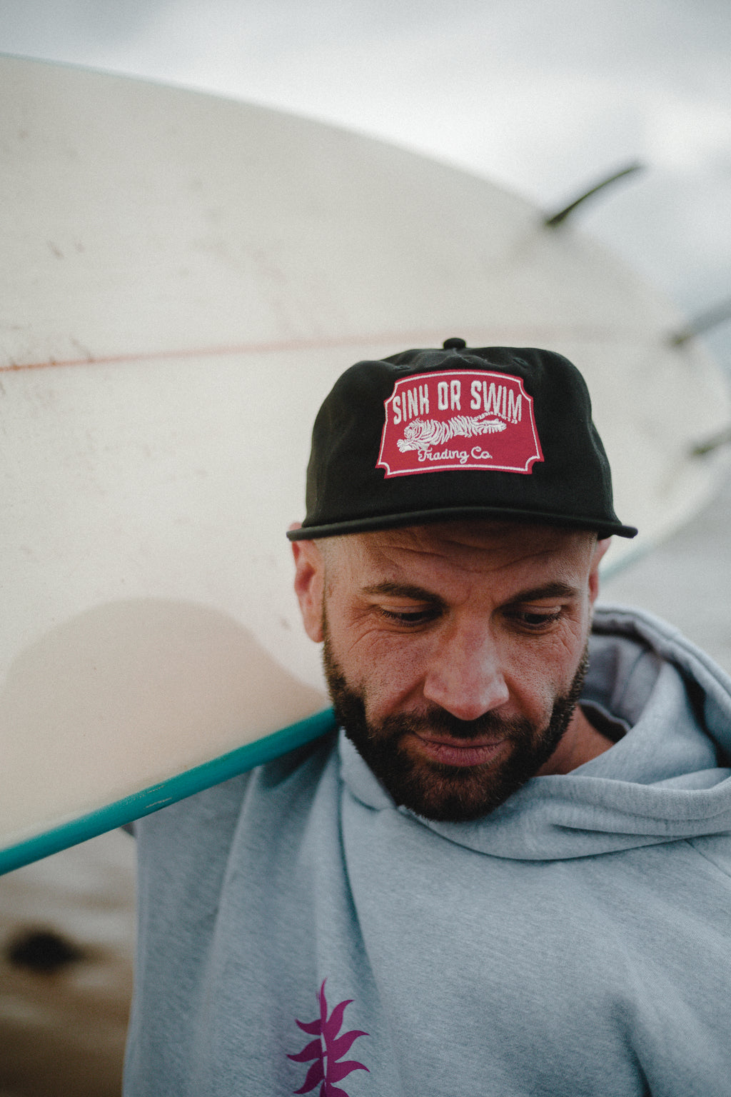 Close-up of a bearded man in the heather grey Neon Sun Wheel hoodie and black Vintage Tiger cap, surfboard on his shoulder