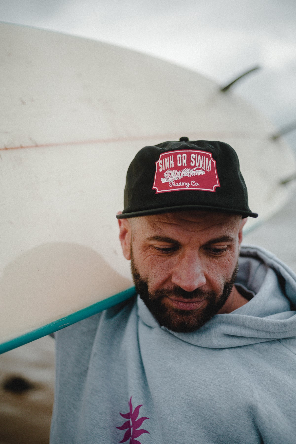 Close-up of a bearded man in the heather grey Neon Sun Wheel hoodie and black Vintage Tiger cap, surfboard on his shoulder