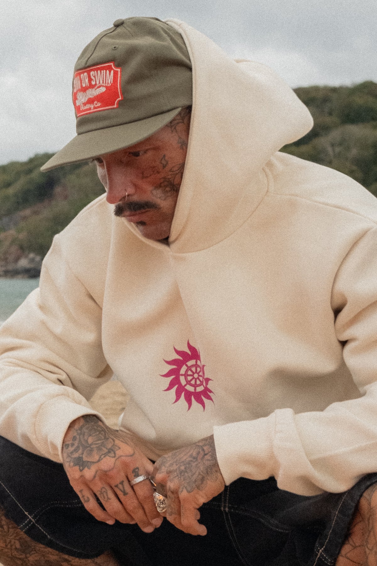 Close-up of the vanilla Neon Sun Wheel hoodie with neon pink sun wheel print, worn with the army green Vintage Tiger cap