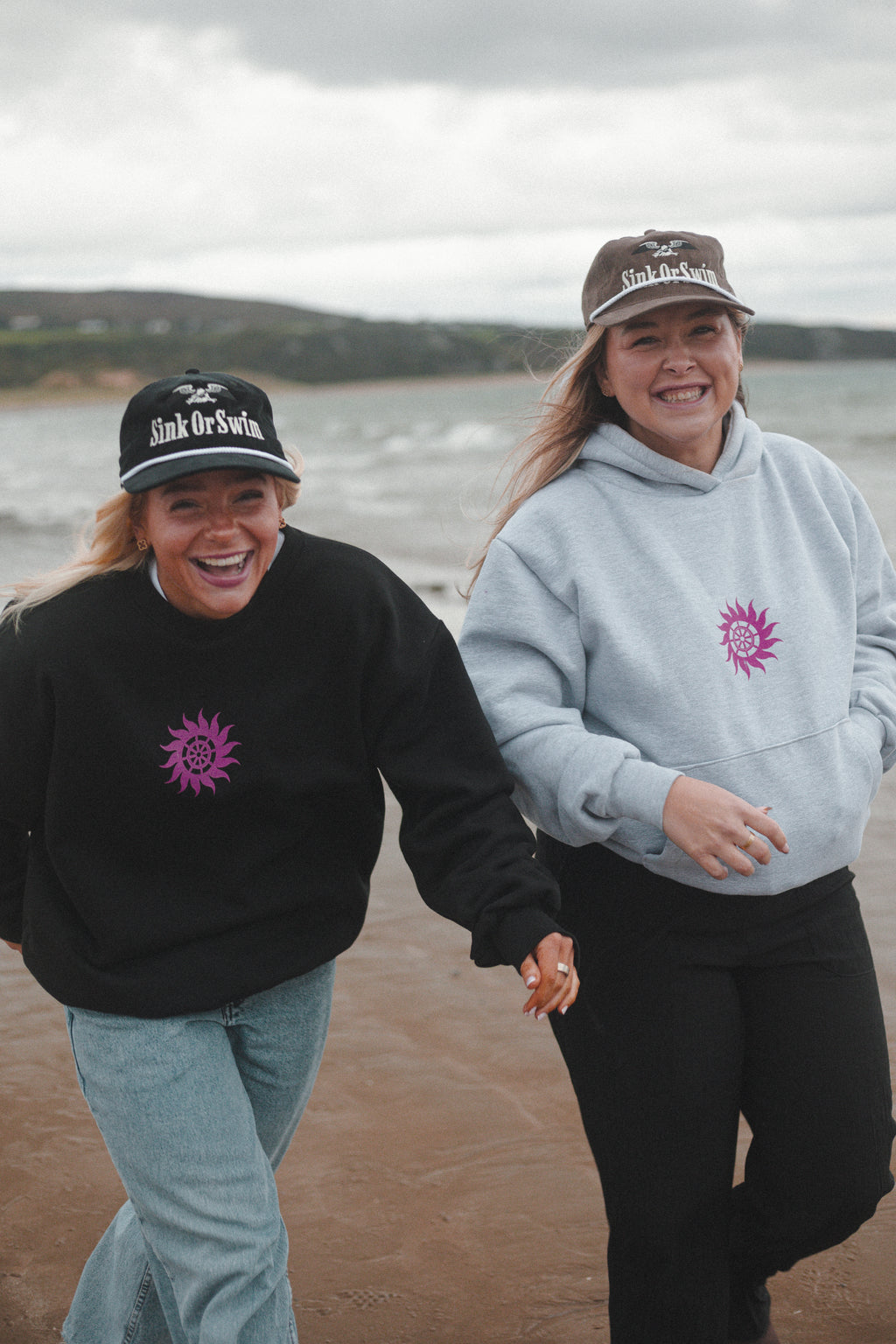 Two women laughing on the beach in a black Neon Sun Wheel sweatshirt and heather grey hoodie with black and brown Eagle caps