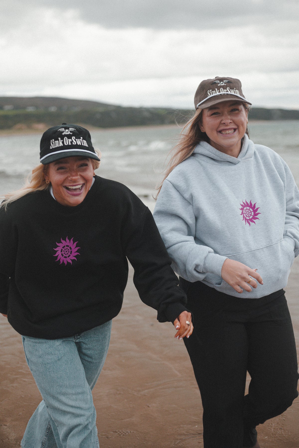 Two women laughing on the beach in a black Neon Sun Wheel sweatshirt and heather grey hoodie with black and brown Eagle caps