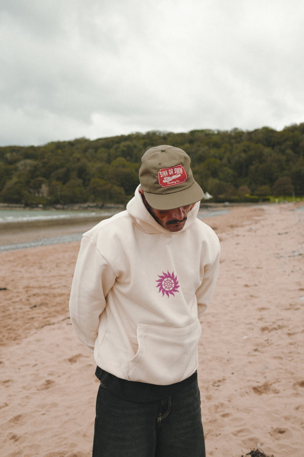 Man in the army green Vintage Tiger 5 panel cap and oversized vanilla Neon Sun Wheel hoodie on a sandy beach