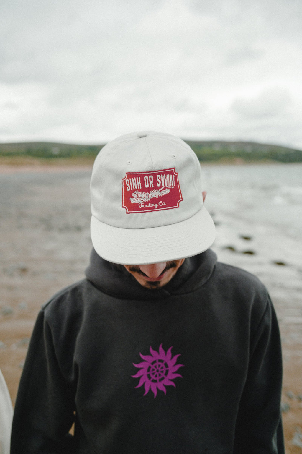 Man wearing the raw canvas Vintage Tiger 5 panel cap and black Neon Sun Wheel hoodie with a neon pink print on the beach