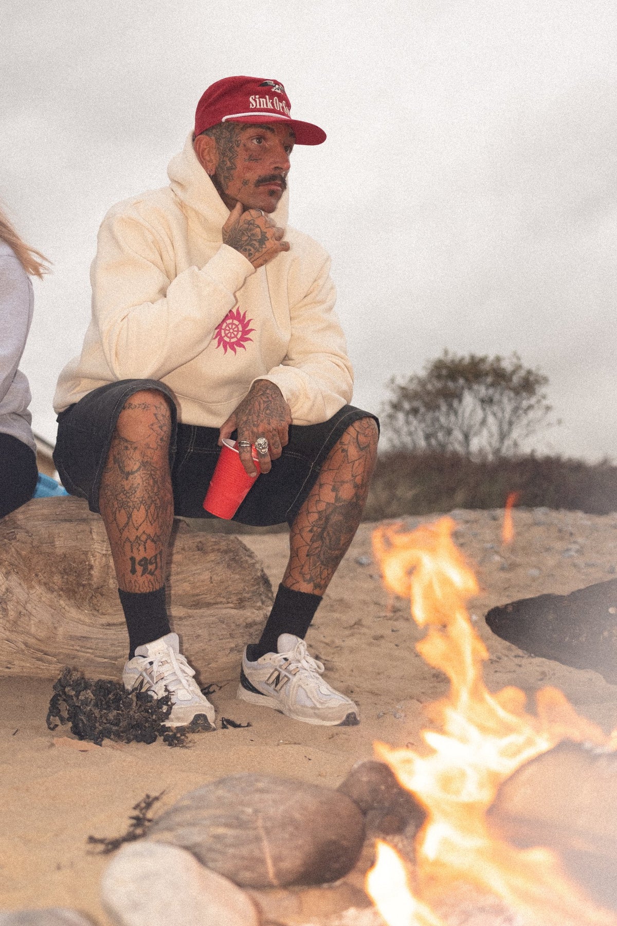 Tattooed man in the red Vintage Eagle cord rope cap and vanilla Neon Sun Wheel hoodie, seated by a beach campfire