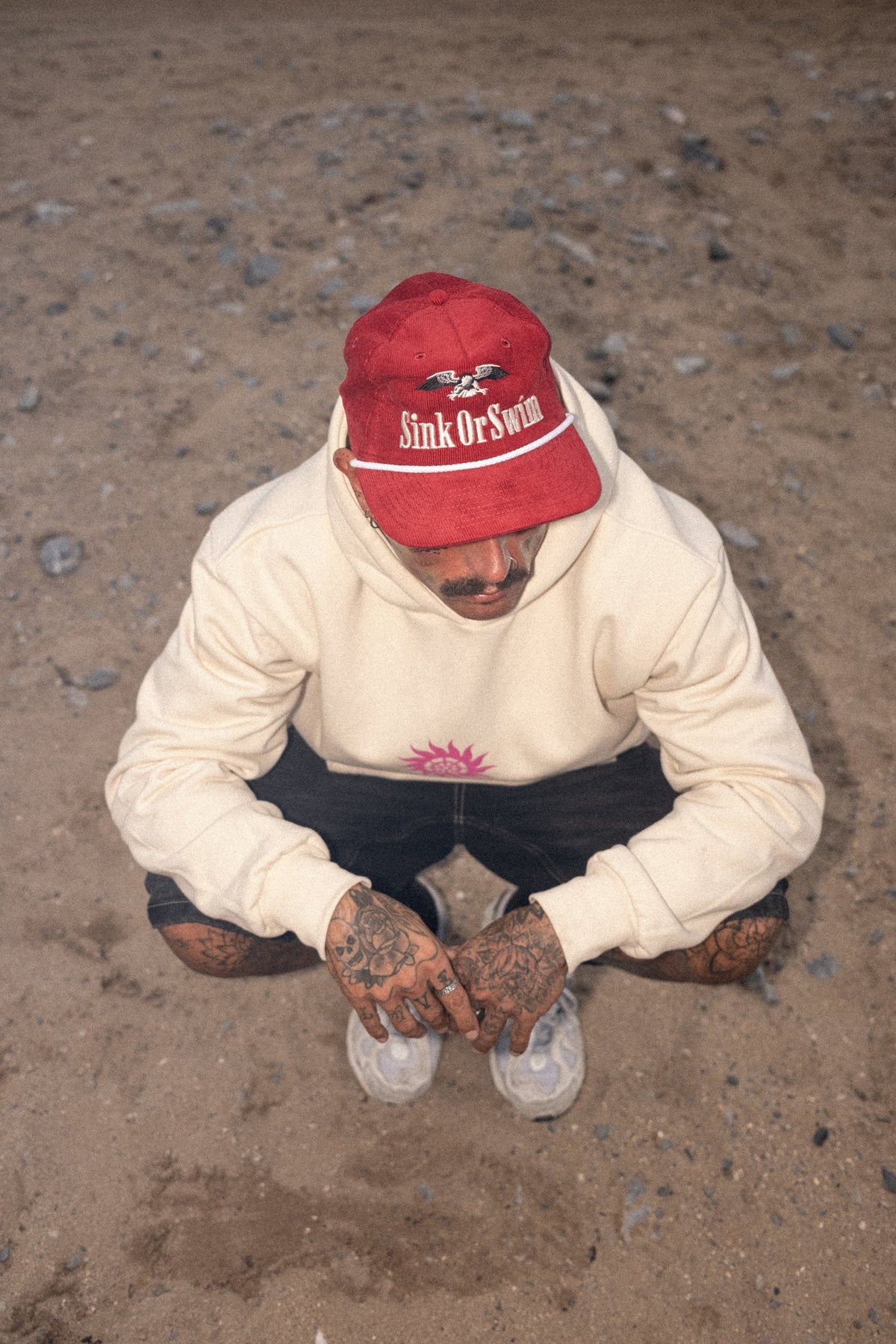 Overhead view of a man crouching on sand in the red corduroy Vintage Eagle rope cap and vanilla Neon Sun Wheel hoodie