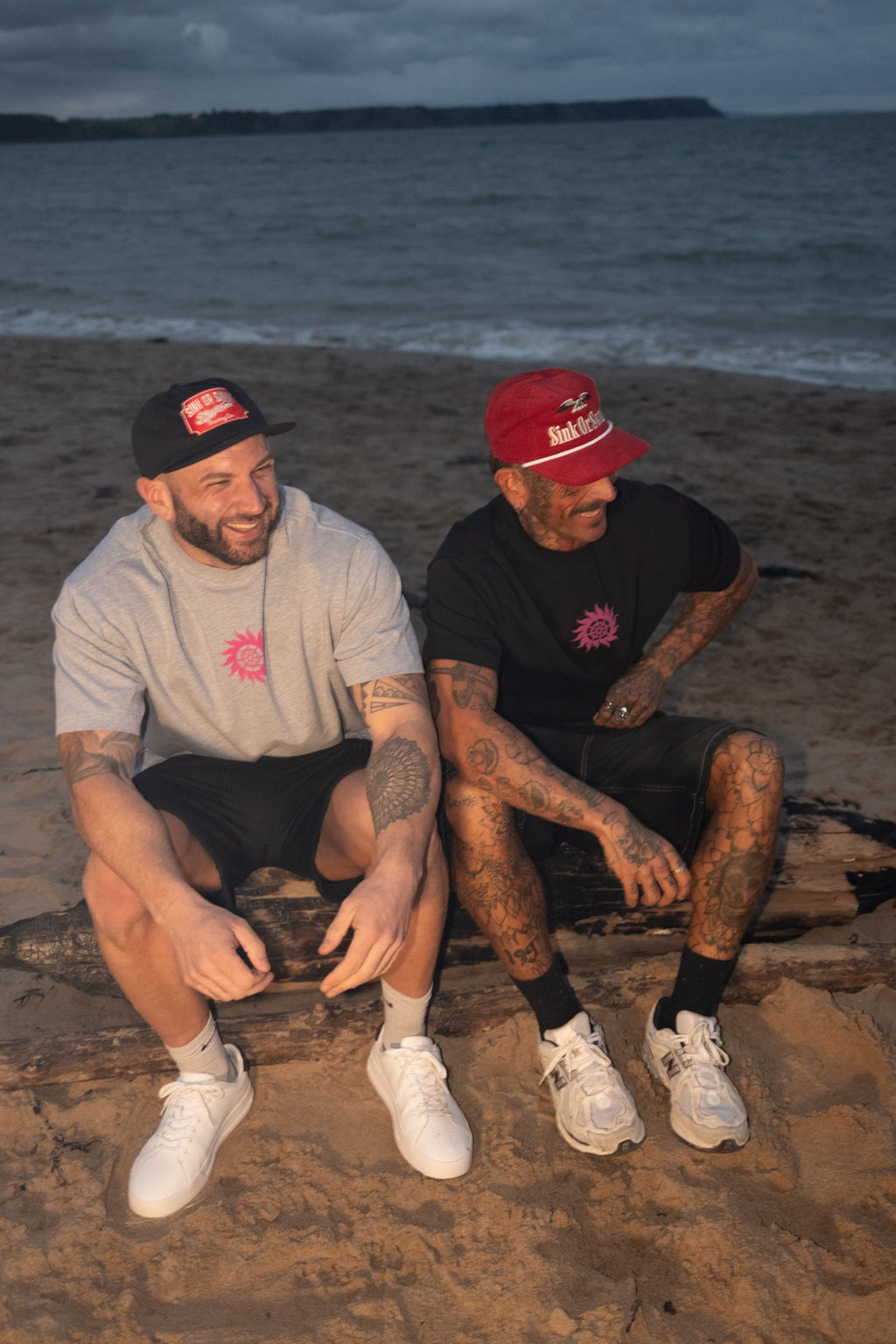 Two men on beach driftwood, one in the red Vintage Eagle cord rope cap and one in the black Vintage Tiger patch cap