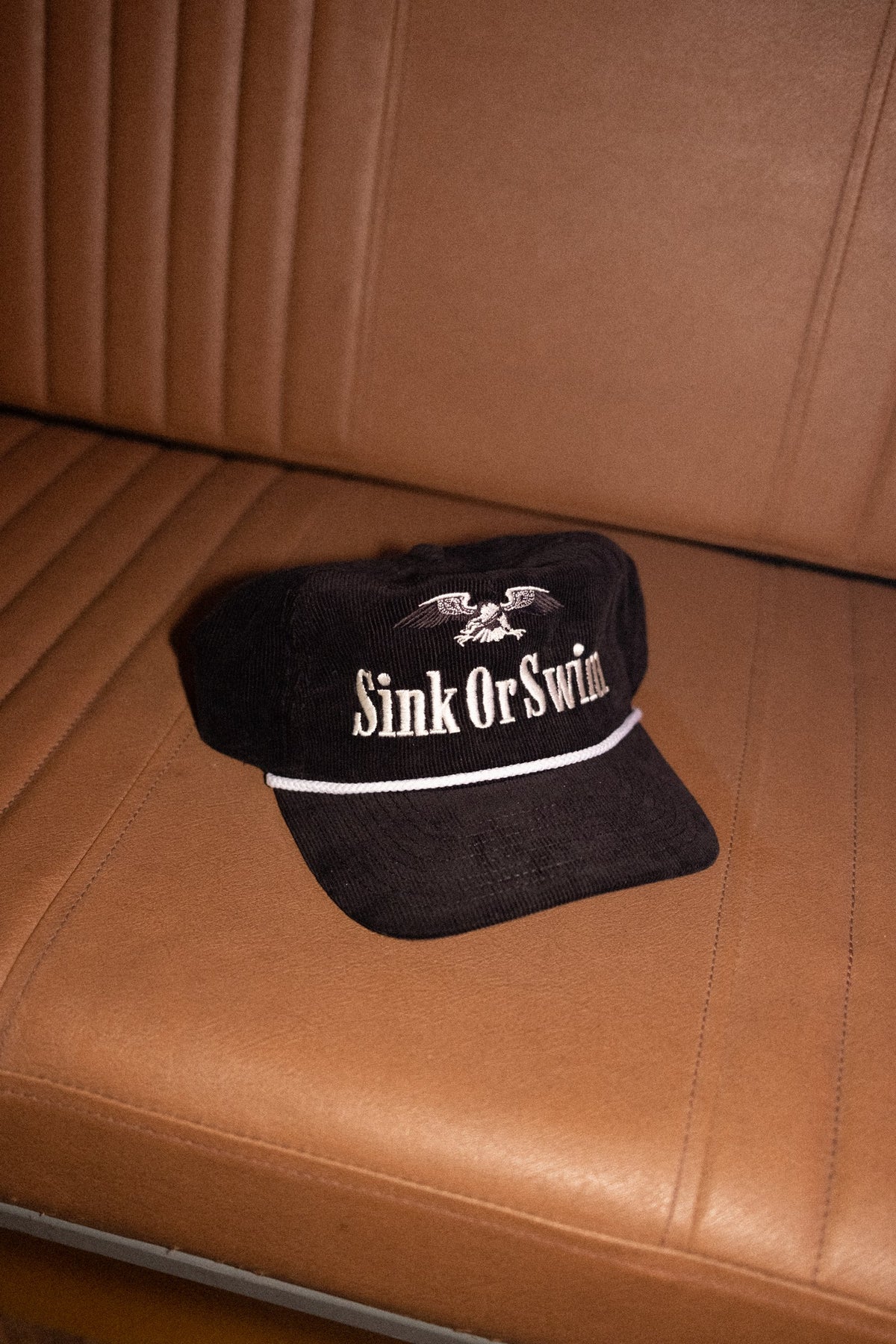 Black corduroy Vintage Eagle rope cap with cream Sink Or Swim script, eagle embroidery and white rope peak, on a leather seat