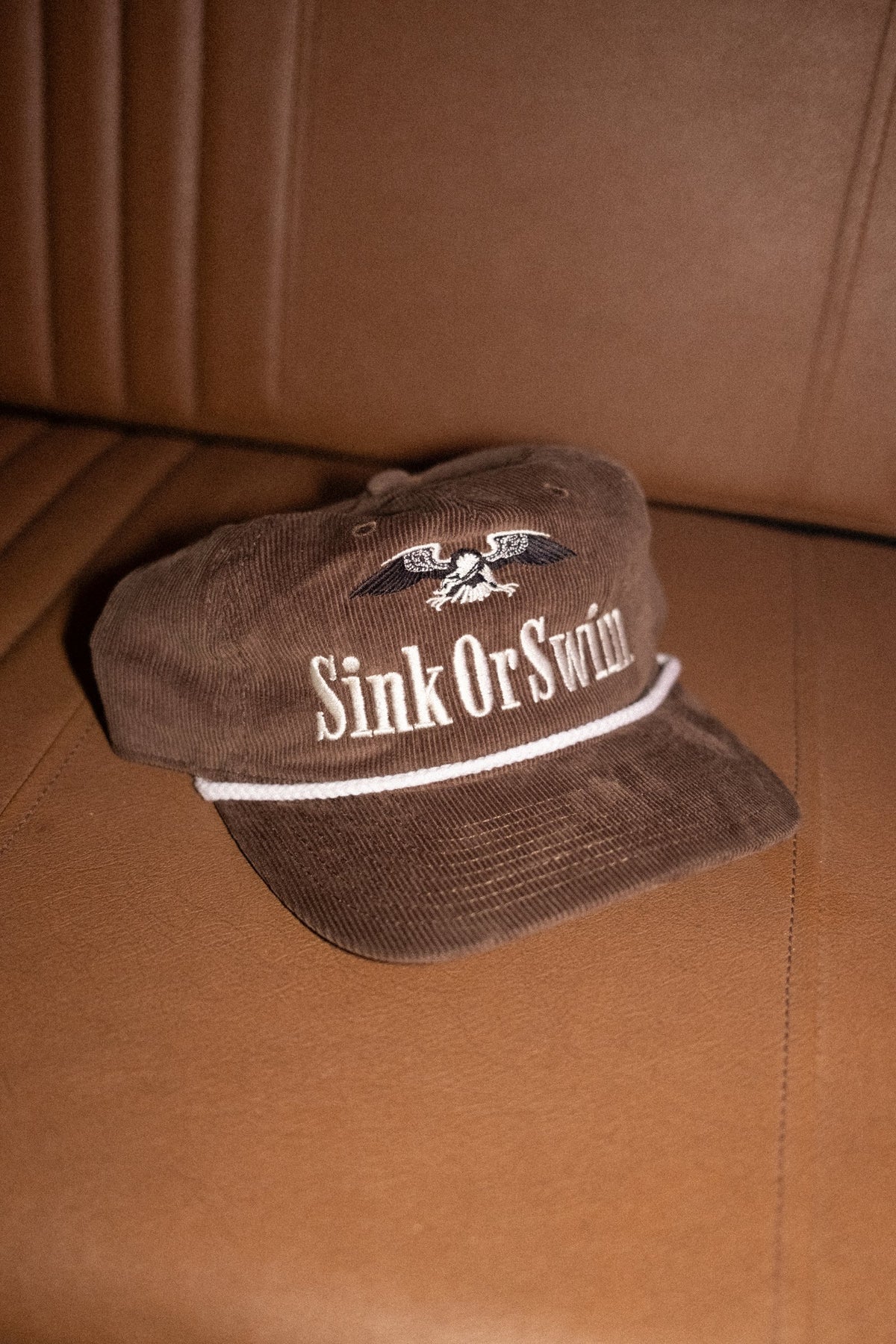 Brown corduroy Vintage Eagle rope cap with cream Sink Or Swim script, eagle embroidery and white rope peak, on a leather seat