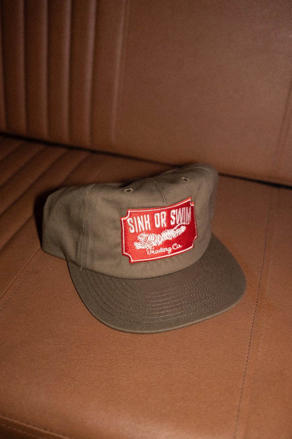Vintage Tiger 5 panel cap in army green cotton canvas with red embroidered Sink Or Swim tiger patch, on a tan leather seat