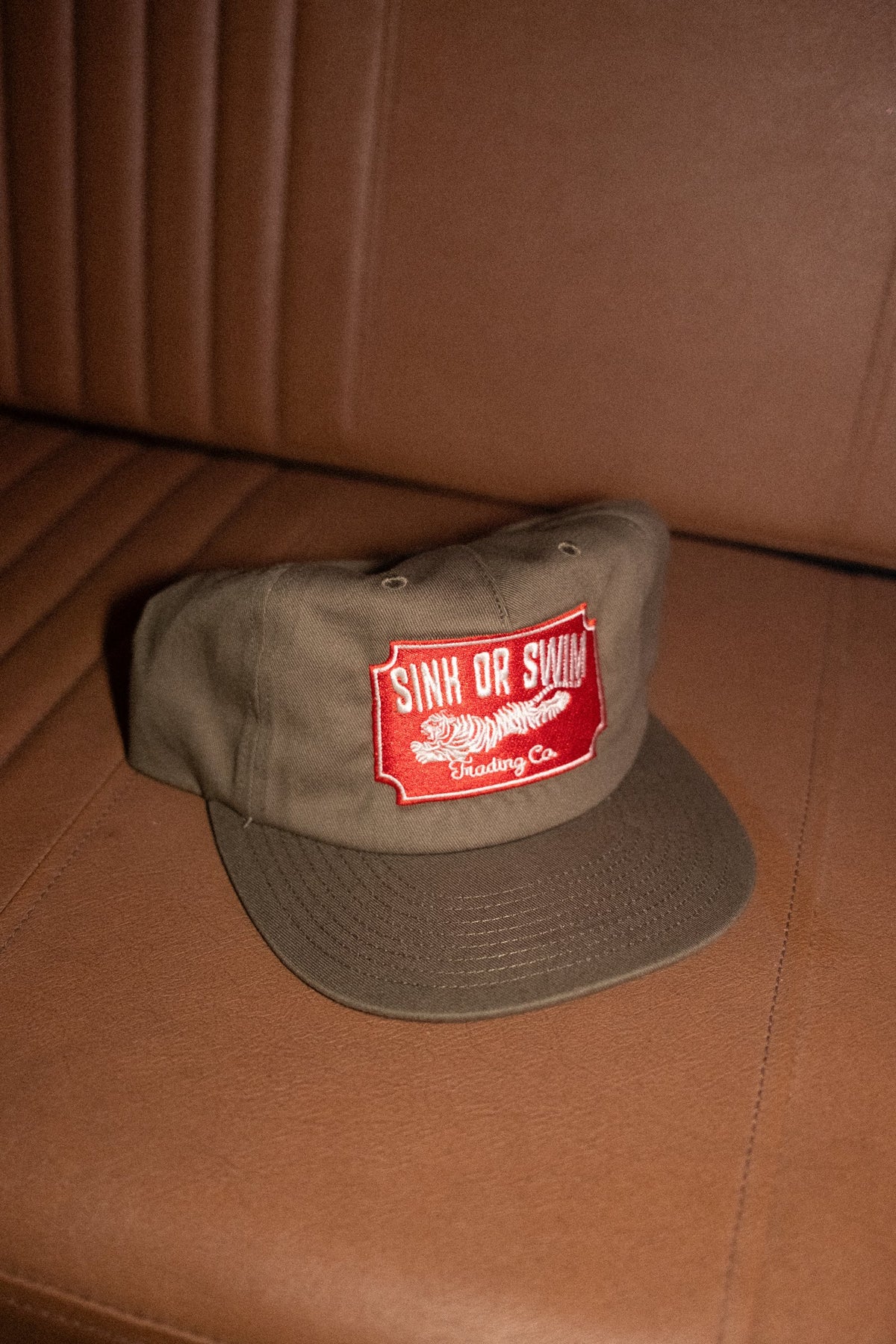 Vintage Tiger 5 panel cap in army green cotton canvas with red embroidered Sink Or Swim tiger patch, on a tan leather seat