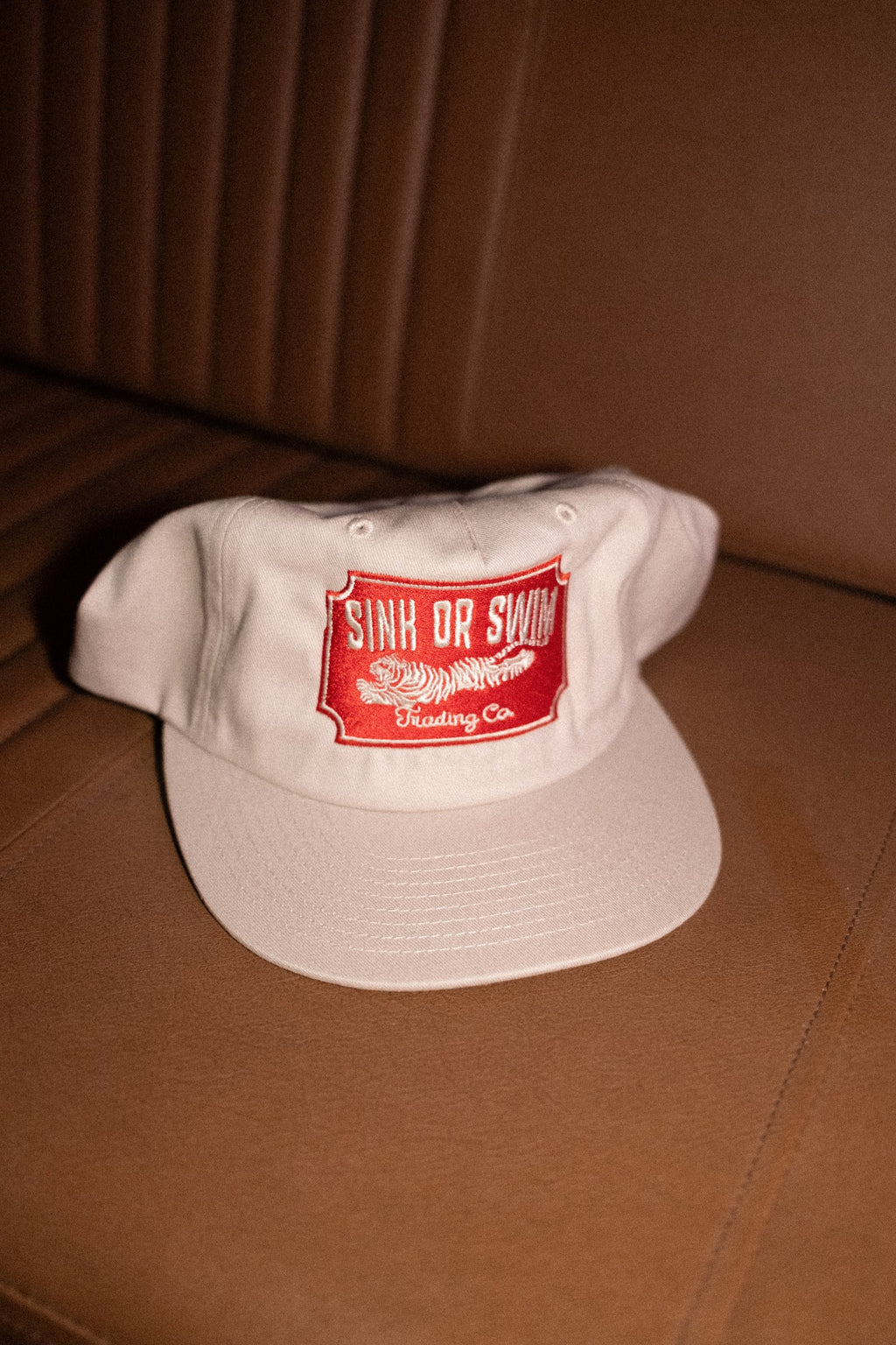 Vintage Tiger 5 panel cap in raw cotton canvas with red embroidered Sink Or Swim tiger patch on a tan leather seat