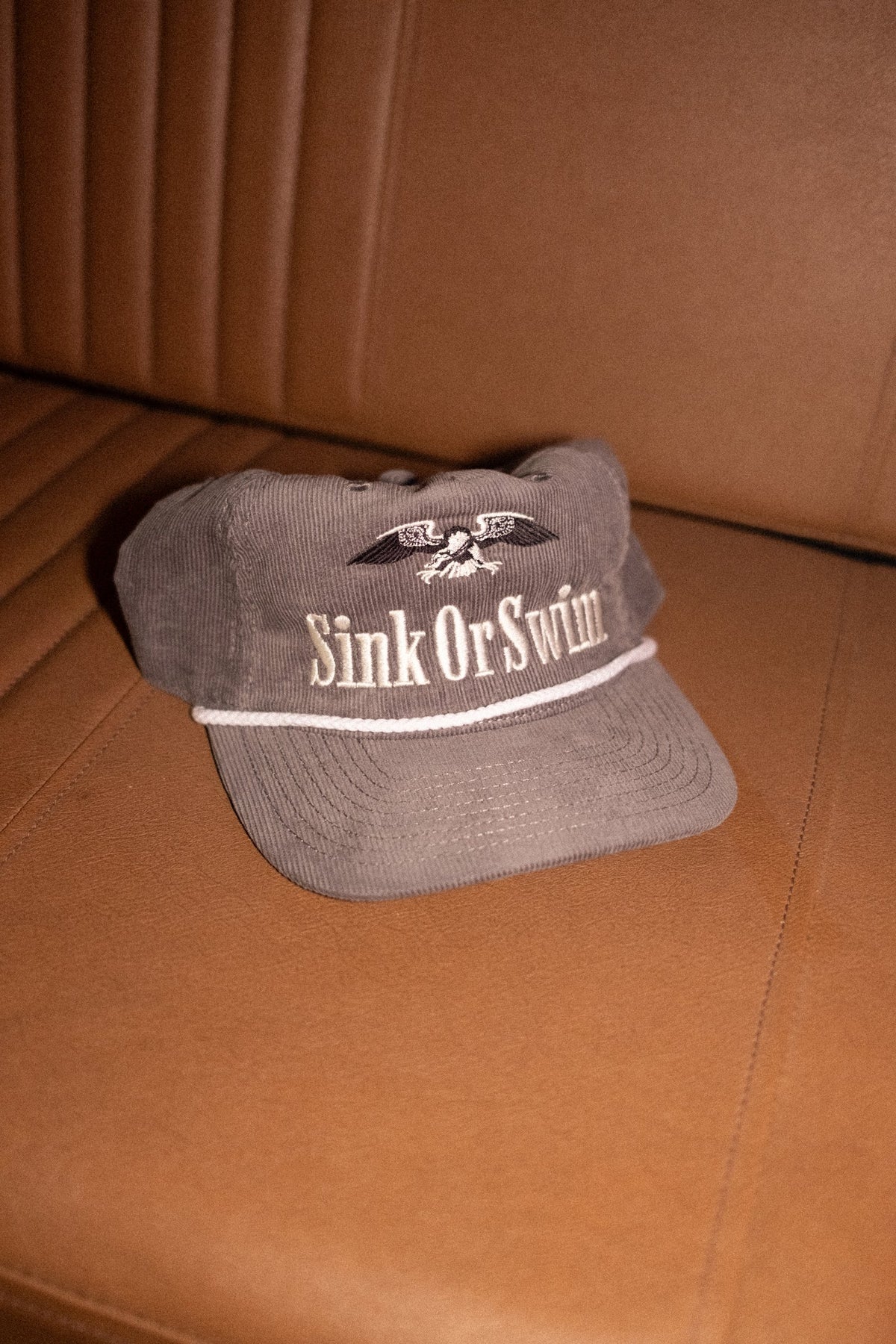 Grey corduroy Vintage Eagle rope cap with cream Sink Or Swim script, eagle embroidery and white rope peak, on a leather seat