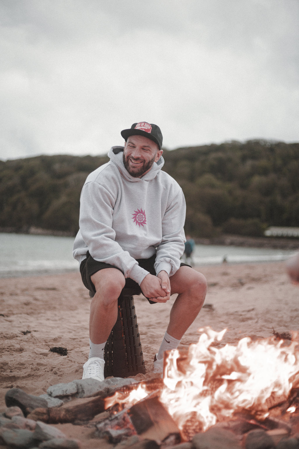Man sitting by a beach campfire in the heather grey Neon Sun Wheel hoodie, black shorts and black Vintage Tiger cap