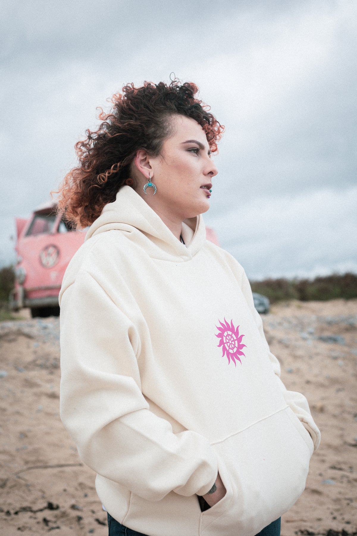 Woman in the vanilla Neon Sun Wheel hoodie, hand in the kangaroo pocket, beside a pink vintage camper van on the beach